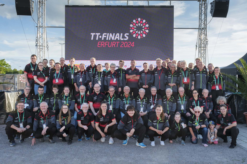 Großes Helferteam der TT-Finals 2024 posiert lächelnd vor einer LED-Bühne im Freien.